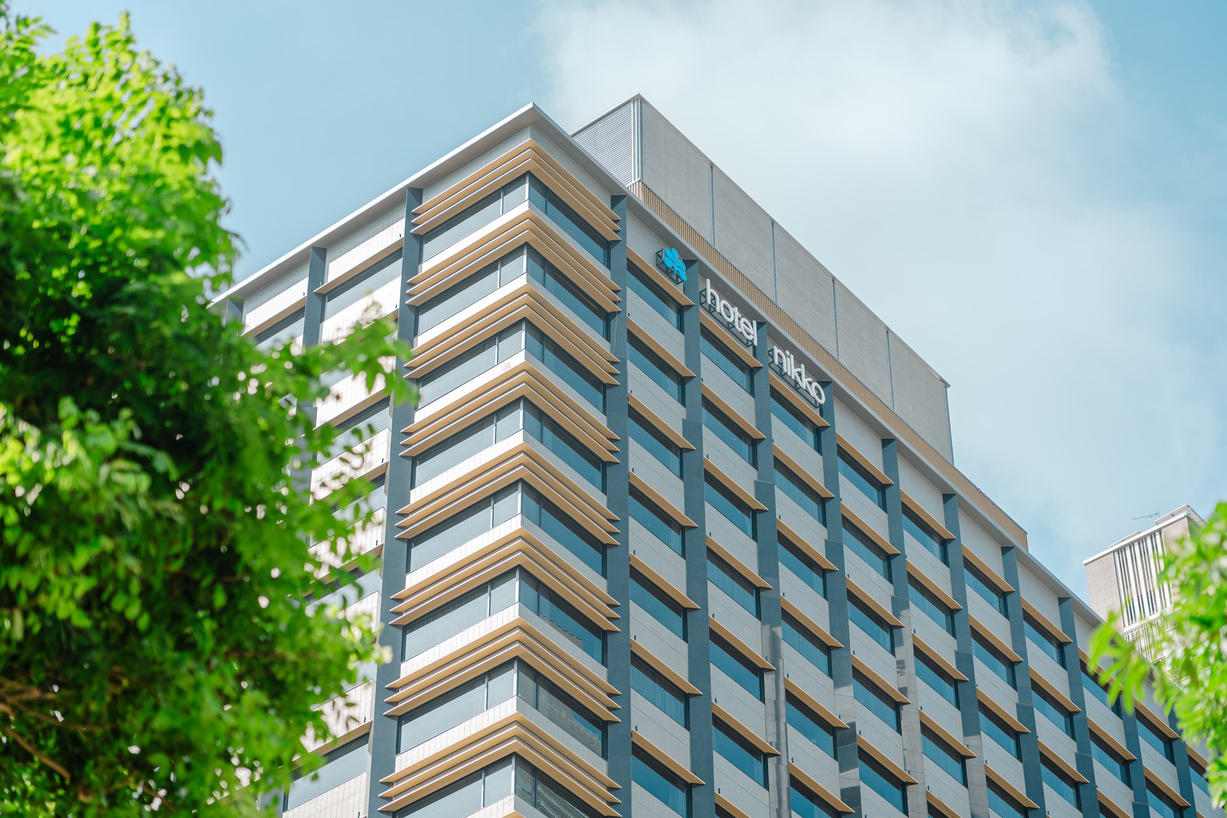 Hotel Nikko Kaohsiung, a modern high-rise with clean lines and uniform balconies, set against a blue sky with leafy green trees in the foreground.