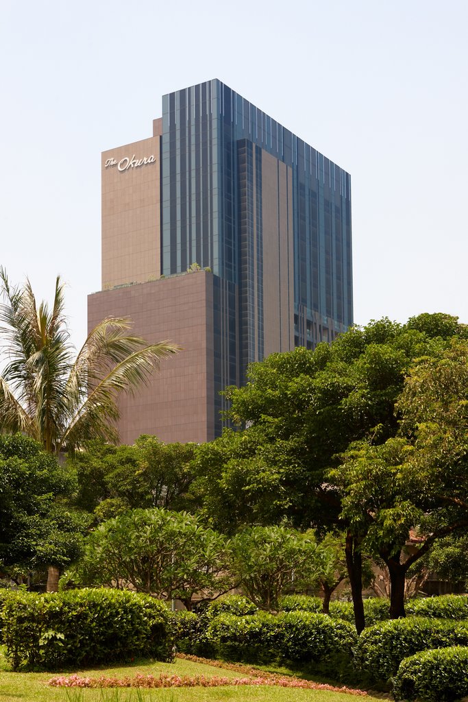 The Okura Prestige Taipei, a tall modern tower with sleek glass and stone exterior, rising above lush tropical greenery.