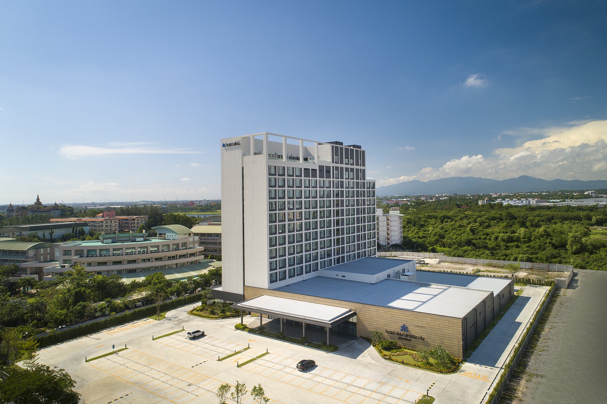 Hotel Nikko Amata City Chonburi, a modern white and gray mid-rise hotel with a flat rooftop and geometric facade, surrounded by greenery and distant mountains under a clear blue sky.