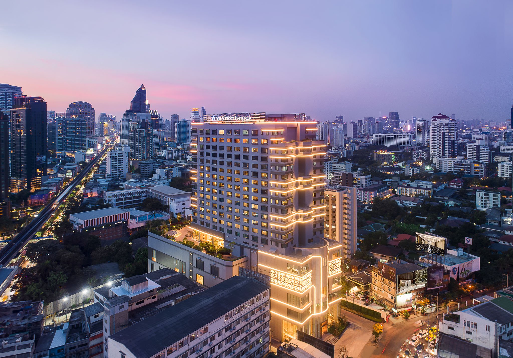 Hotel Nikko Bangkok, a modern high-rise building illuminated with warm linear lights, set in a vibrant cityscape at dusk with surrounding skyscrapers and a lively urban atmosphere.
