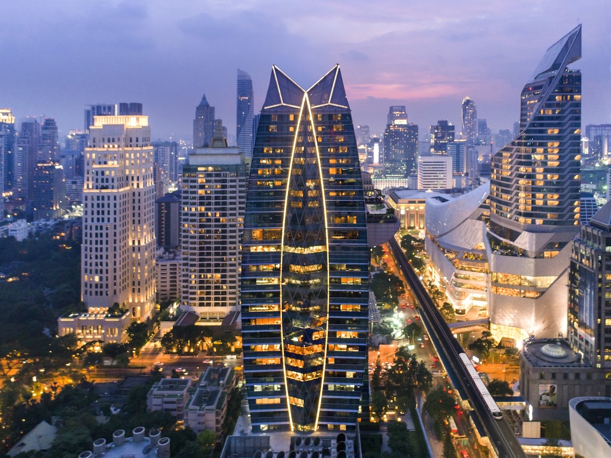 he Okura Prestige Bangkok, a distinctive V-shaped skyscraper with illuminated edges, standing out in a vibrant city skyline at dusk, surrounded by high-rise buildings and an elevated train line.