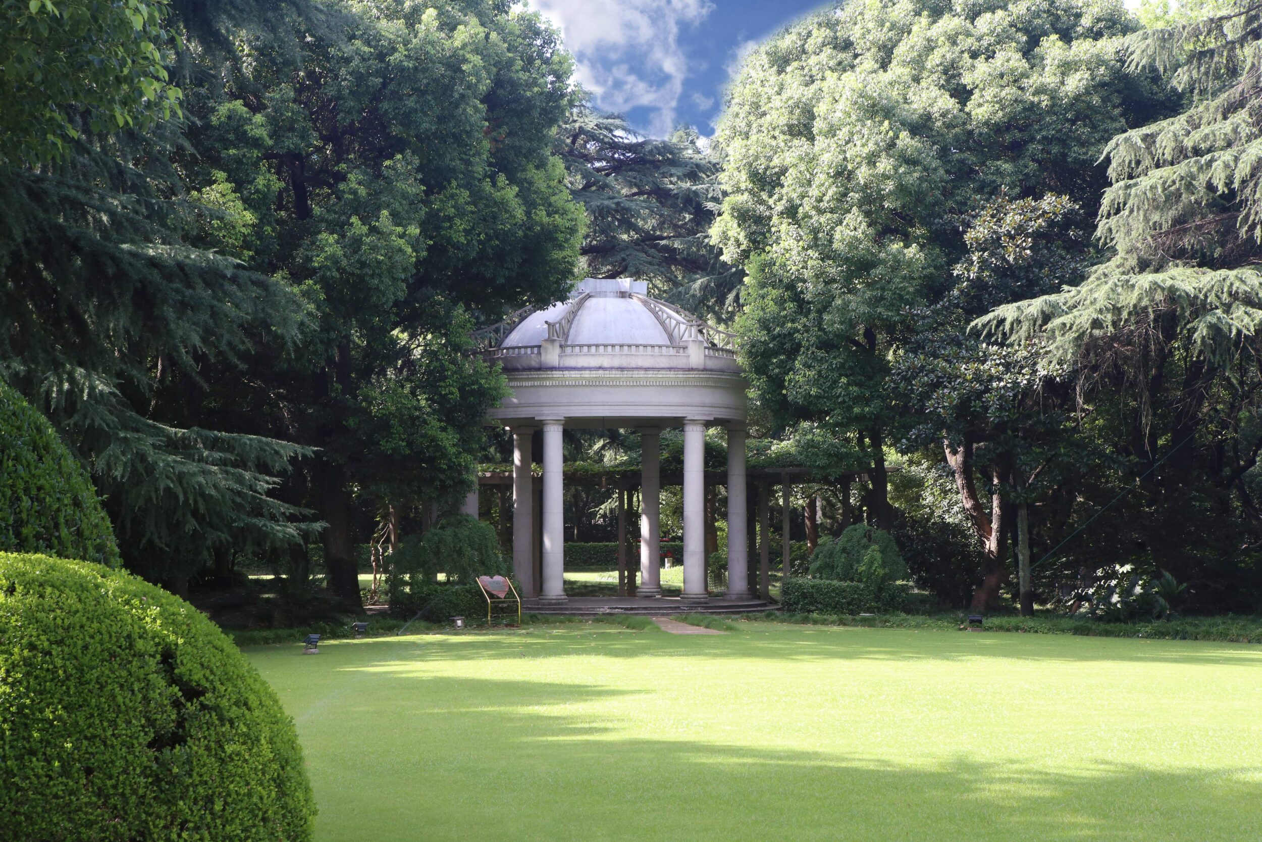 A wide shot of a lush green lawn leading to a classical, circular white gazebo with columns and a dome roof, nestled within a dense grove of tall, mature trees.