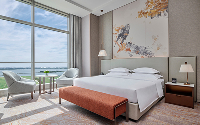 A bright hotel bedroom with a large bed, featuring an orange upholstered bench at the foot. Floor-to-ceiling windows offer a water view. Two light armchairs sit by a round table near the window.