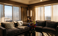 A warmly lit corner living room with a dark grey L-shaped sofa and chevron pillows. Large windows with sheer and heavy gold curtains offer a bright city view. A colorful flower bouquet sits on a coffee table in the center.
