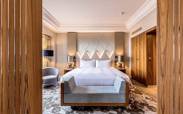 A luxurious hotel bedroom with a large bed featuring white linens and a tufted, beige diamond-patterned headboard. The room includes a grey upholstered bench at the foot of the bed and matching bedside tables with lamps. The foreground is framed by dark wood paneling with silver vertical inlays.