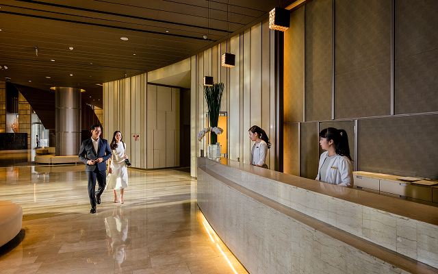 A wide shot of a luxurious hotel lobby with a long, light marble reception desk. Two women in white hotel uniforms are standing behind the desk. A well-dressed couple is walking toward the desk on the left. The lobby features gold-toned lighting and wall paneling.