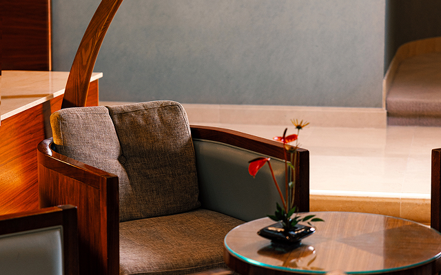 A close-up of a dark wood armchair with beige upholstery, next to a glass table with a red flower in a vase.