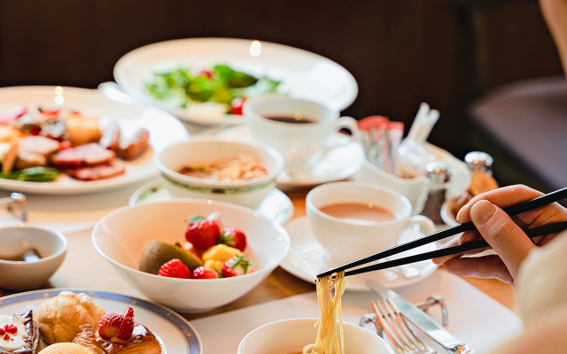 A hand uses black chopsticks to eat noodles, surrounded by a large table spread of fruit and breakfast dishes.