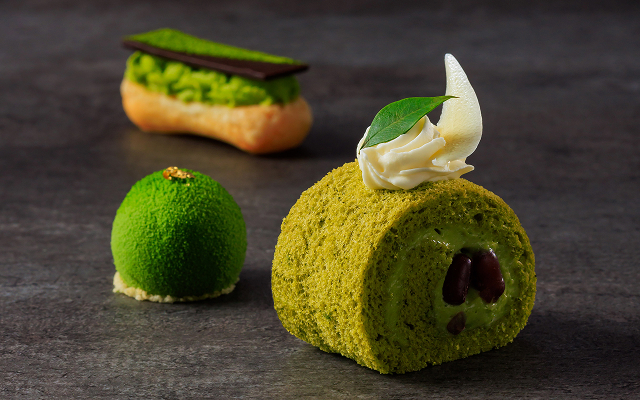 A dark image of three matcha desserts on a grey surface, including a green Swiss roll with red bean, a small green dome mousse, and a matcha eclair.