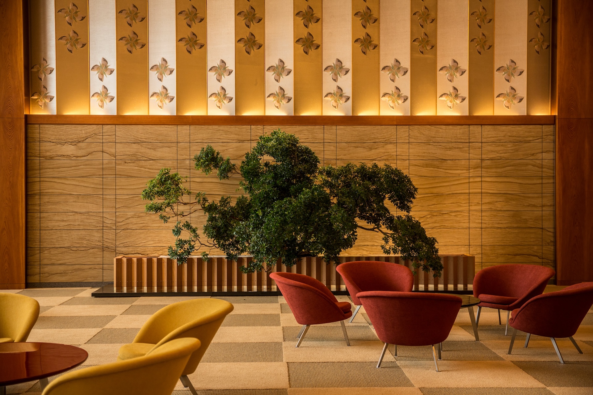 stylish hotel lobby or lounge with a checkered light beige floor. The room features a grouping of modern chairs in mustard yellow and burnt orange. The wall is decorated with horizontal wood paneling below a row of vertical panels featuring gold and cream stripes