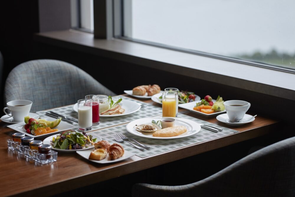 A table set for two with a large breakfast spread next to a window. The meal includes plates of fresh fruit, a plate with an oyster and a pastry, croissants and bread, milk and red juice, side salads, and two cups of coffee.