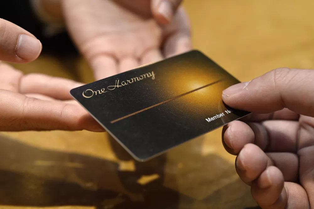Two hands are exchanging a dark "One Harmony" membership card with a central gold light effect.