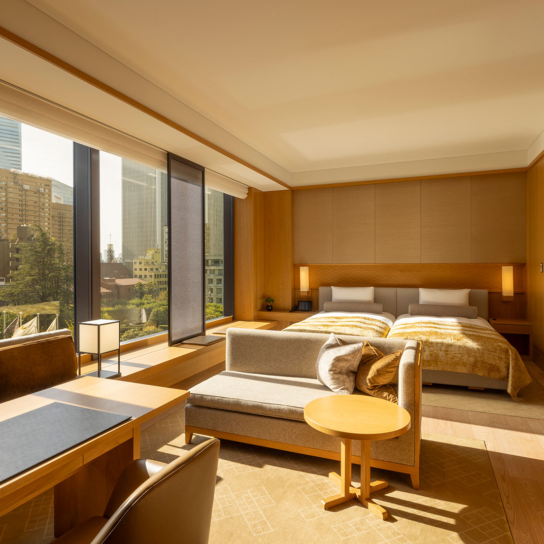 A spacious and bright hotel room suite, featuring a sitting area with a sofa, a small round table, and a work desk in the foreground. Two single beds with yellow and white covers are in the background. Large windows overlook a sunny cityscape and nature.