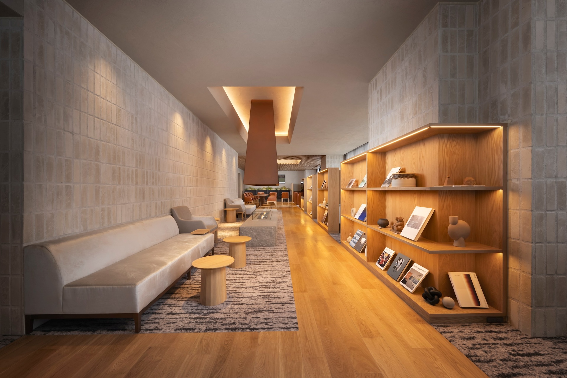 A modern, long lounge or library with light wood floors, textured beige walls, and a large bookshelf. A long, light sofa sits on a rug on the left, facing an illuminated display shelf on the right.
