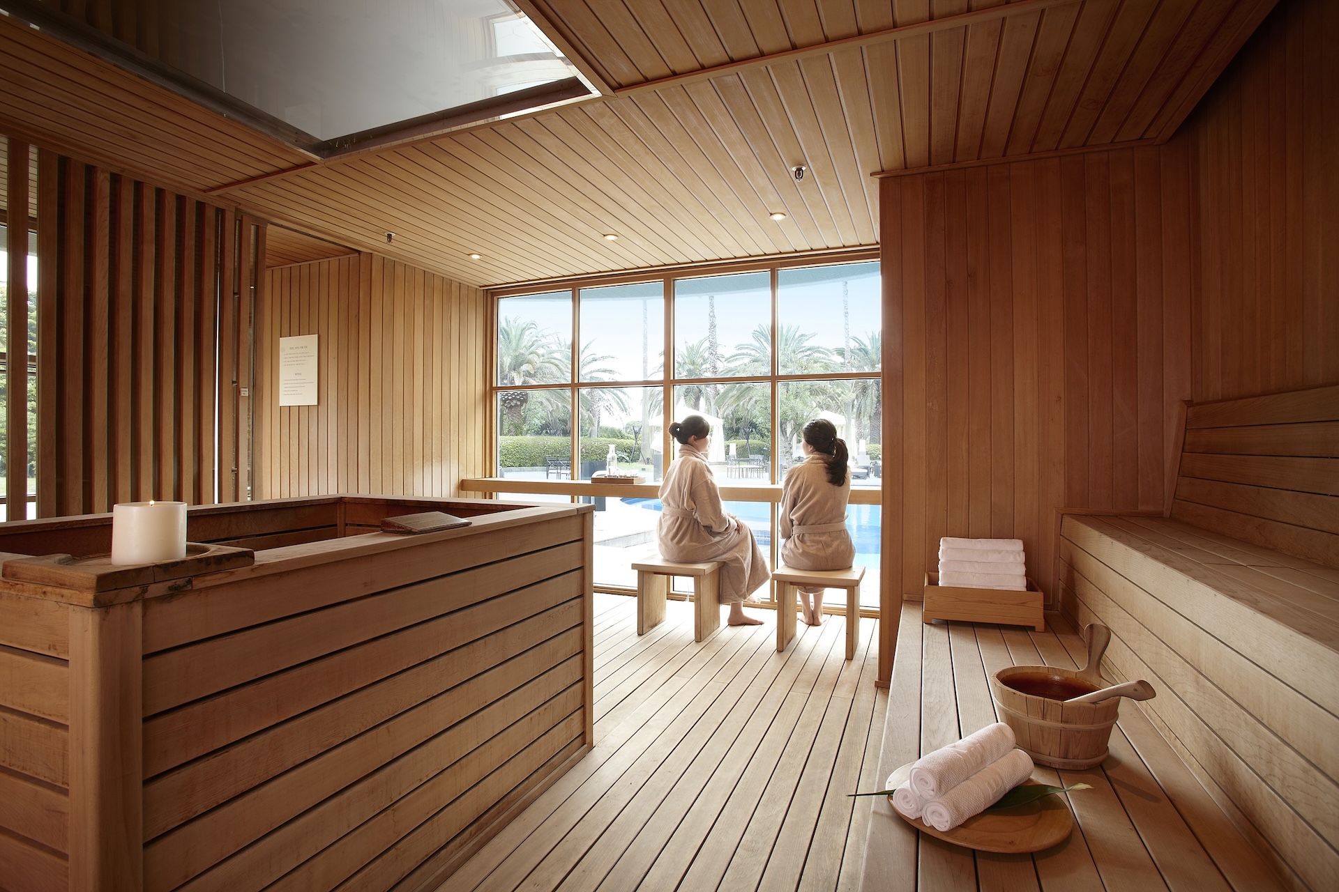 A wooden sauna or relaxation room with two women in robes looking out a large window at a pool and palm trees.