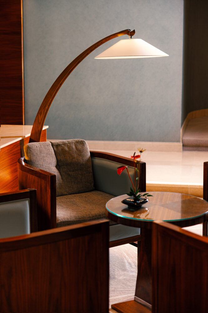A comfortable, modern armchair with dark wood framing and gray upholstery, illuminated by a tall, curved floor lamp. A small glass-top table holds a flower arrangement.