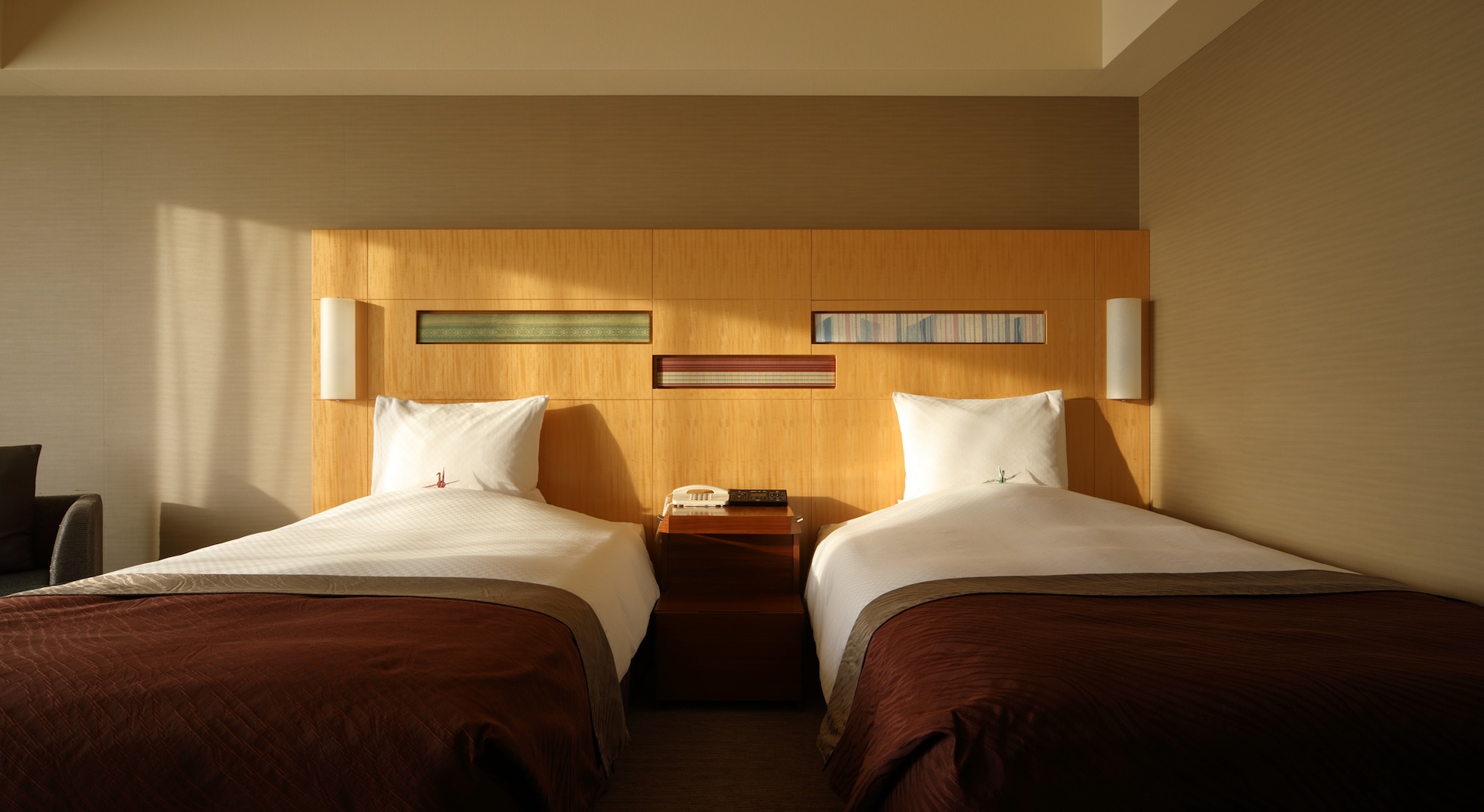 A warm, modern twin hotel room with two single beds, each with white bedding and a dark brown blanket. A light wood headboard with integrated lighting and small decorative panels connects the beds, and sunlight streams in from the left, casting shadows across the room.