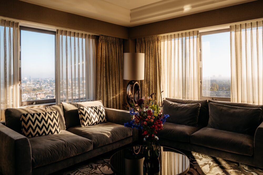 A luxurious, high-floor hotel living room or suite lounge with plush gray couches and zigzag patterned pillows. Large windows, draped with sheer and velvet curtains, overlook a city view bathed in soft sunlight.