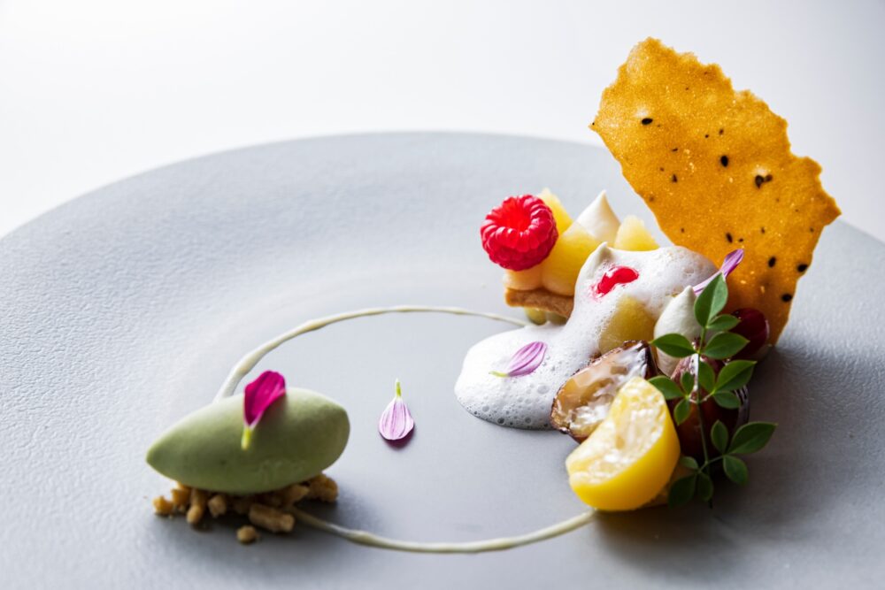 A gourmet plated dessert on a gray ceramic dish, featuring a scoop of green sorbet on crumbles, a raspberry on a small tart, meringue foam, citrus slices, and a large, savory-looking crisp, garnished with small pink flower petals.