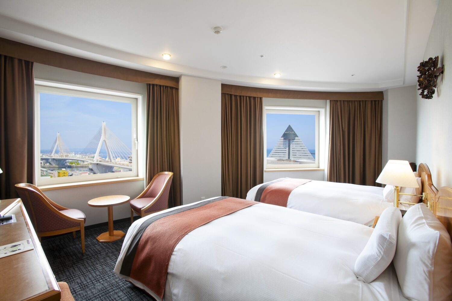 A bright twin hotel room with two large windows. The left window shows a view of a large suspension bridge, while the right window frames a distinctive pyramid-shaped building against the sea. The room has two single beds, brown curtains, and a small sitting area.