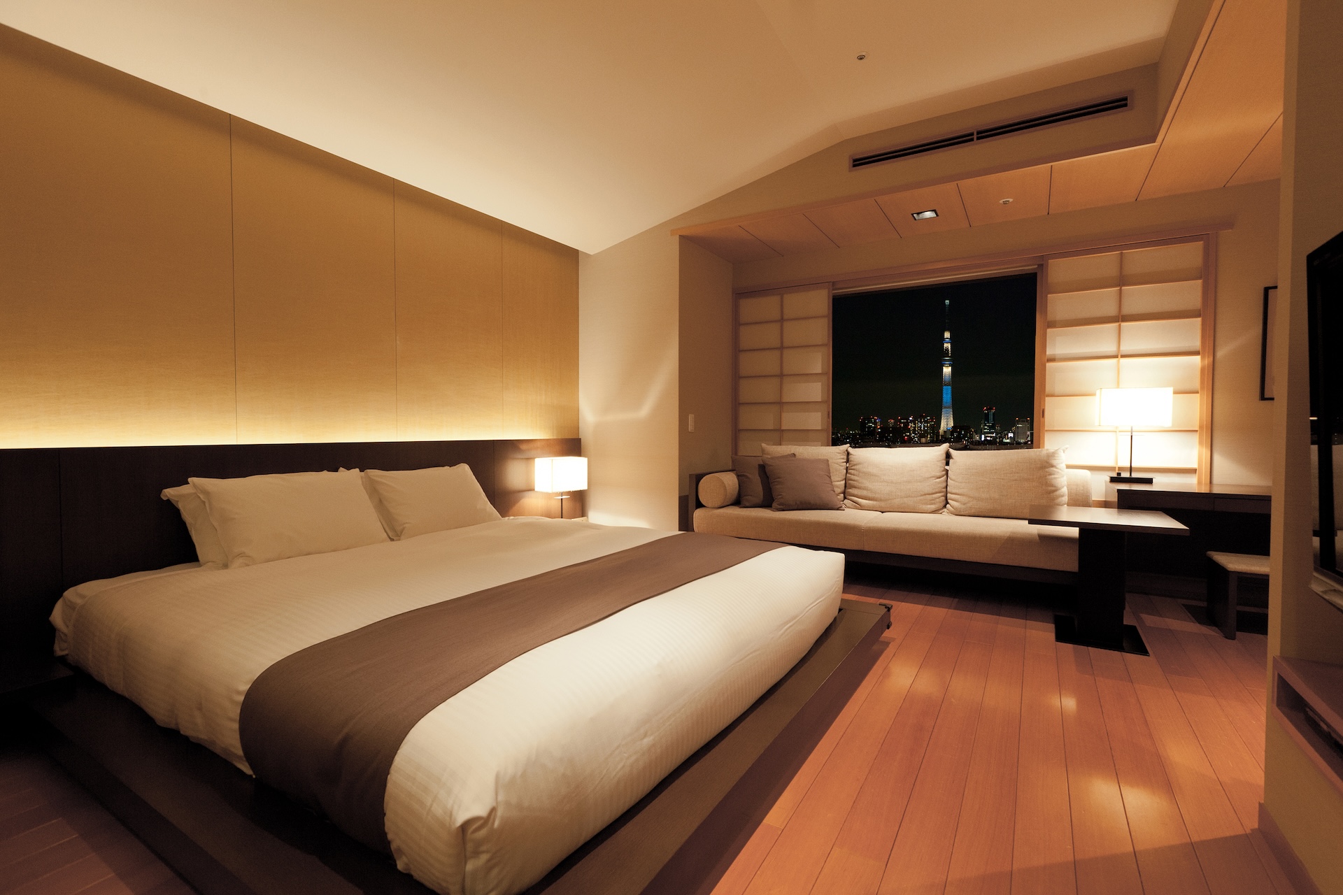 A modern, Japanese-inspired hotel room at night, featuring a large bed with a brown bed runner and hardwood floors. A recessed window area with shoji screens holds a long sofa overlooking a brightly lit city skyline, including the illuminated Tokyo Skytree.