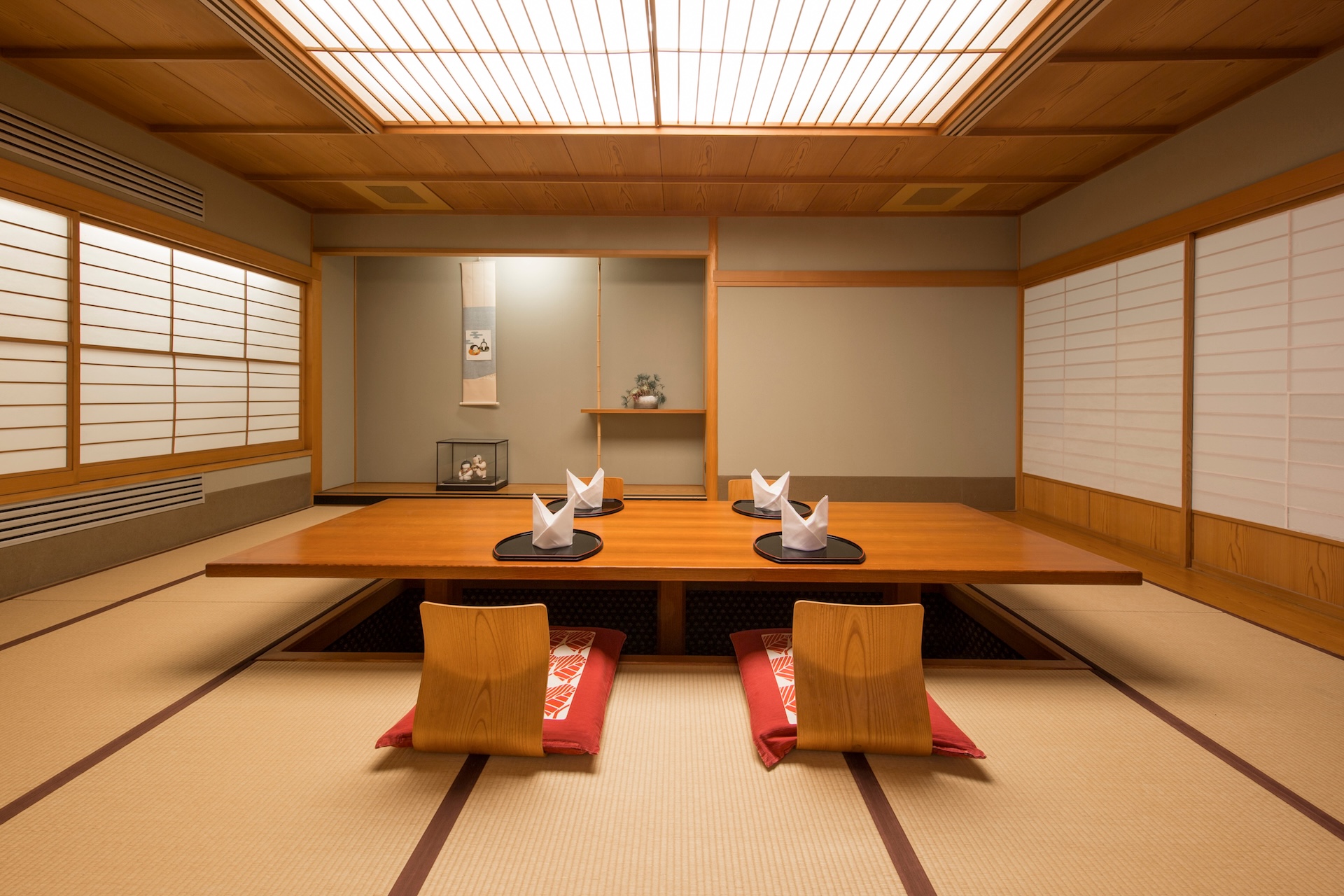 A traditional Japanese dining room (washitsu) featuring a low, wooden table with recessed seating (horigotatsu style). The room has tatami flooring, shoji screen windows, a wooden lattice ceiling with soft lighting, and minimal decor in the alcove (tokonoma). Two place settings with folded napkins are visible.