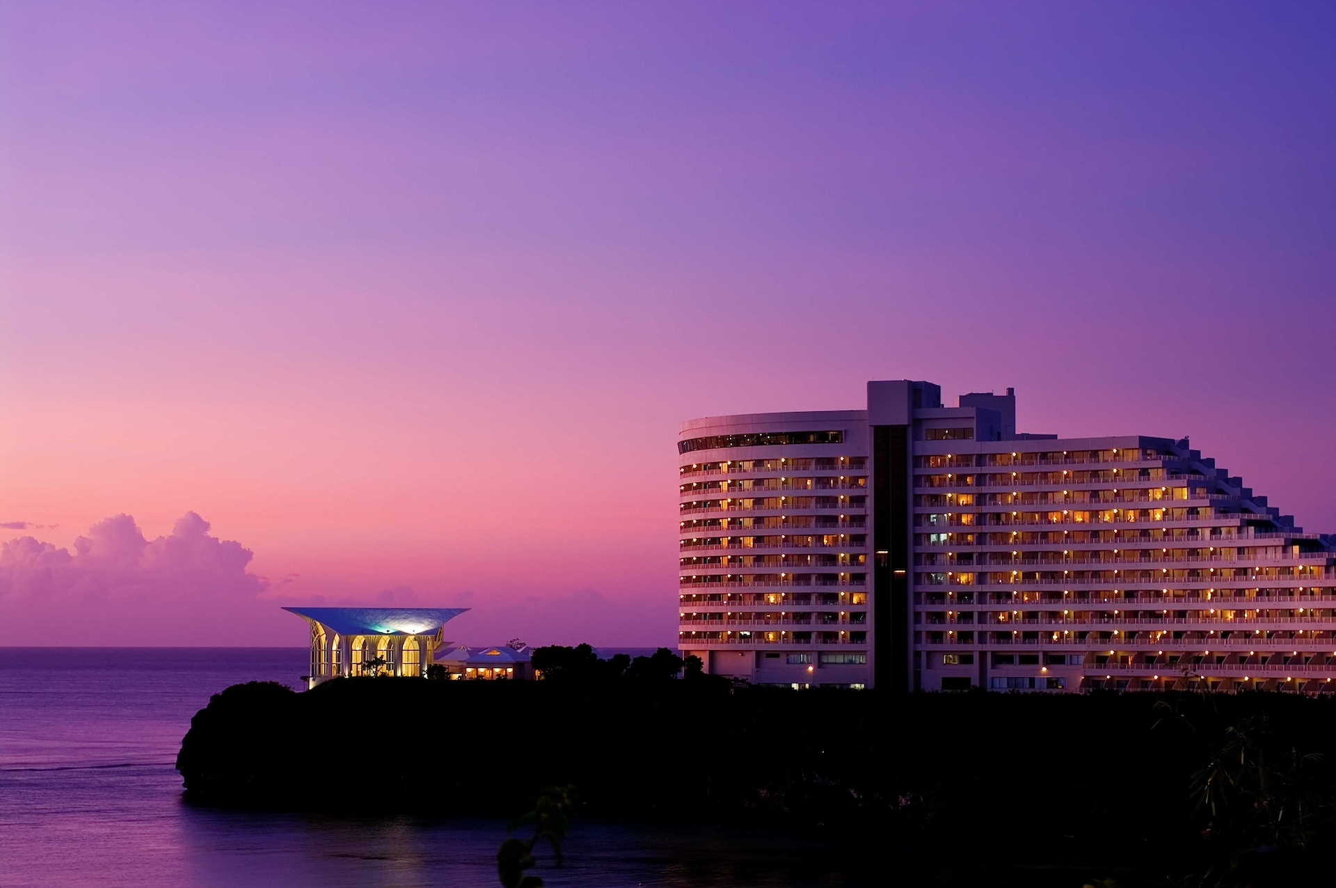 resort hotel complex, featuring a tiered, terraced main building and a smaller, round auxiliary building, set against a dramatic deep purple and pink sunset over the ocean. The hotel windows are warmly lit.