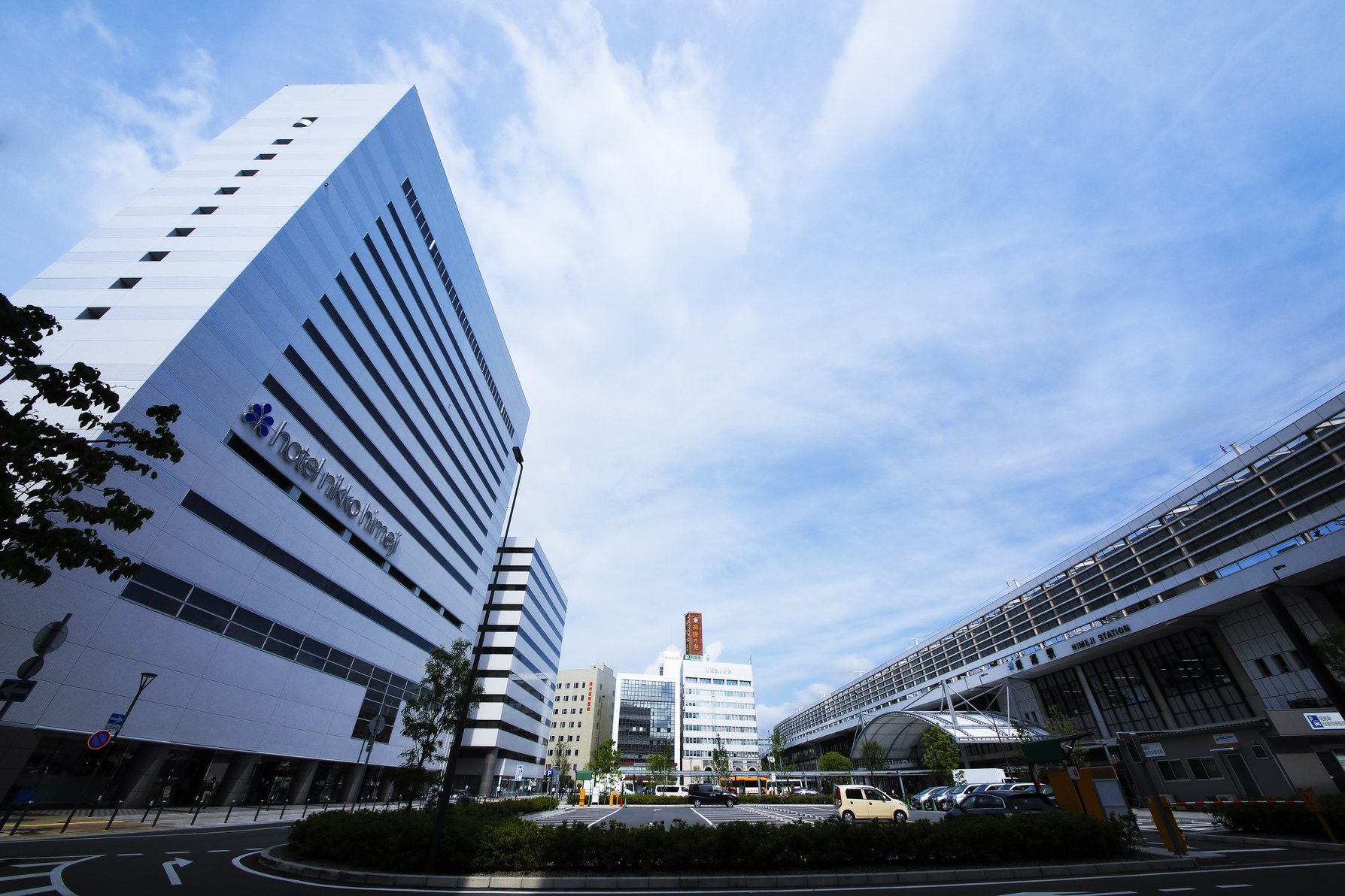 Hotel Nikko Himeji with its modern white facade beside Himeji Station under a bright blue sky.