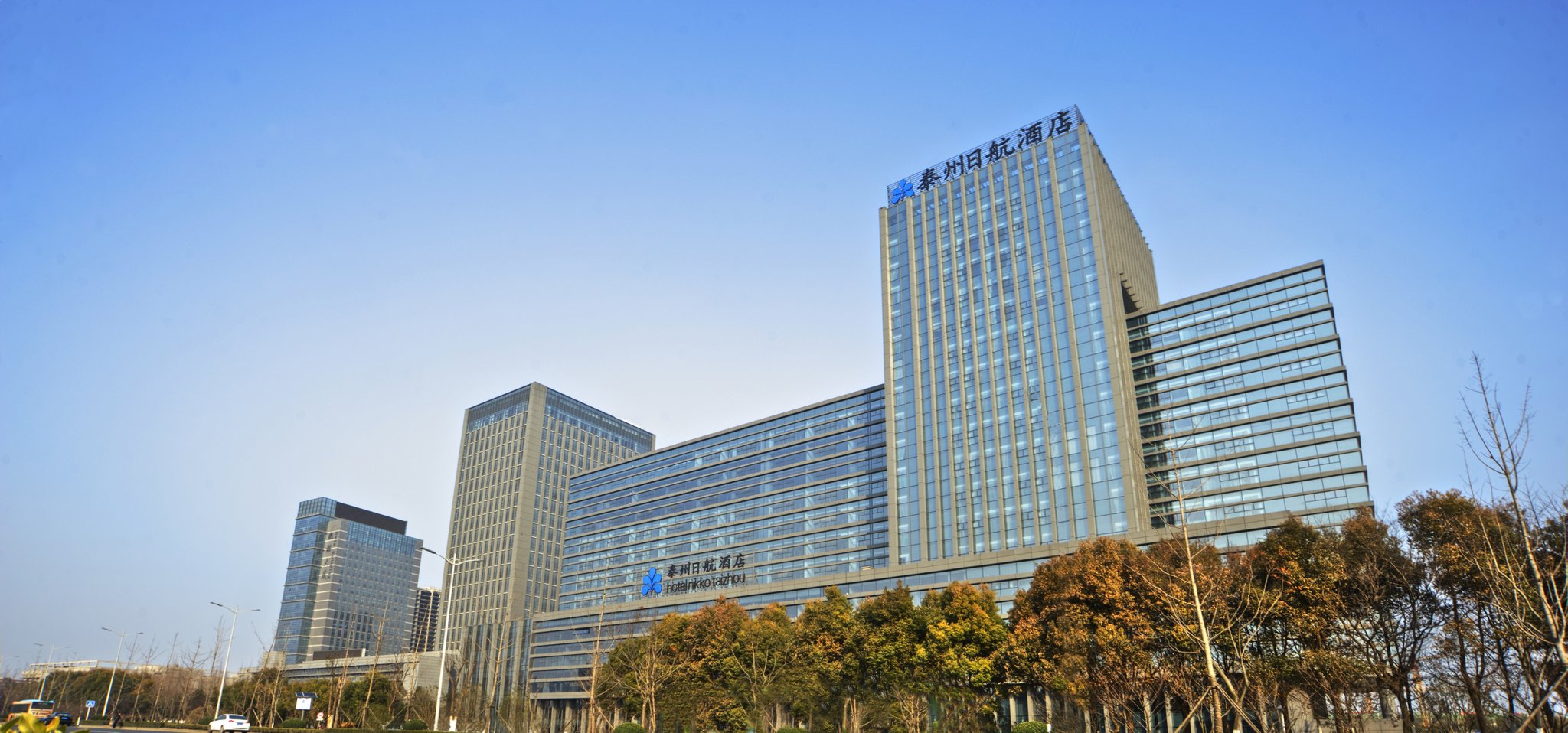 Hotel Nikko Taizhou, a sleek glass tower with modern architecture, rises above tree-lined streets under a clear blue sky.