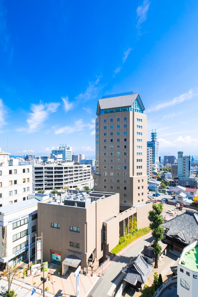 Hotel JAL City Nagano, a tall beige tower with a distinctive gabled roof, stands prominently among urban buildings under a bright blue sky.