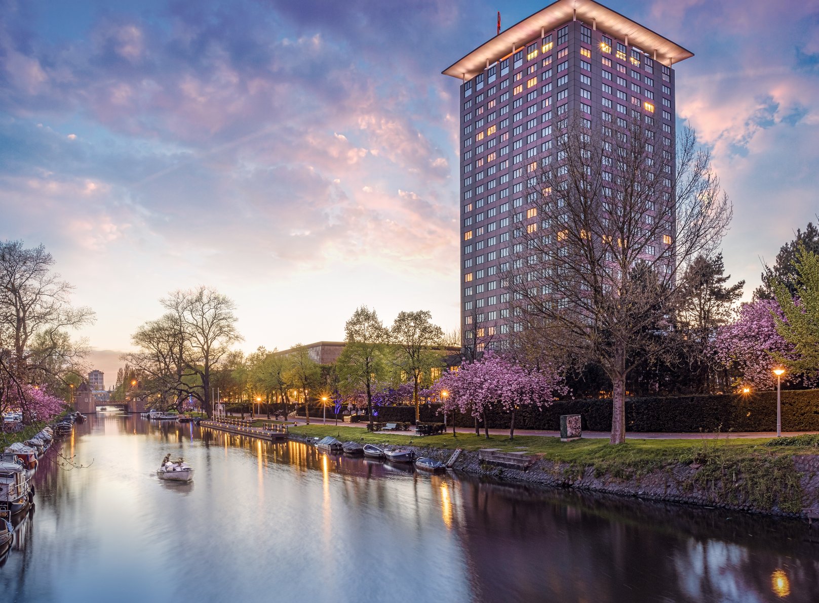 Hotel Okura Amsterdam, a tall riverside tower lit at dusk, rises beside a canal lined with cherry blossom trees and small boats.