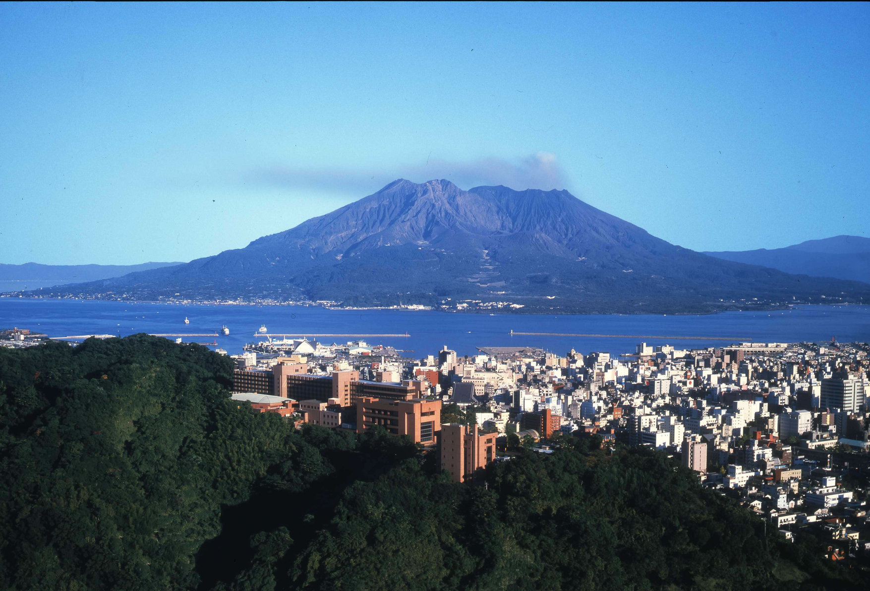 Cityscape of Kagoshima with SHIROYAMA HOTEL in foreground and Sakurajima volcano in background.
