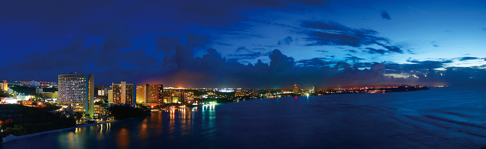 Coastal city skyline at night with hotels and ocean reflections.