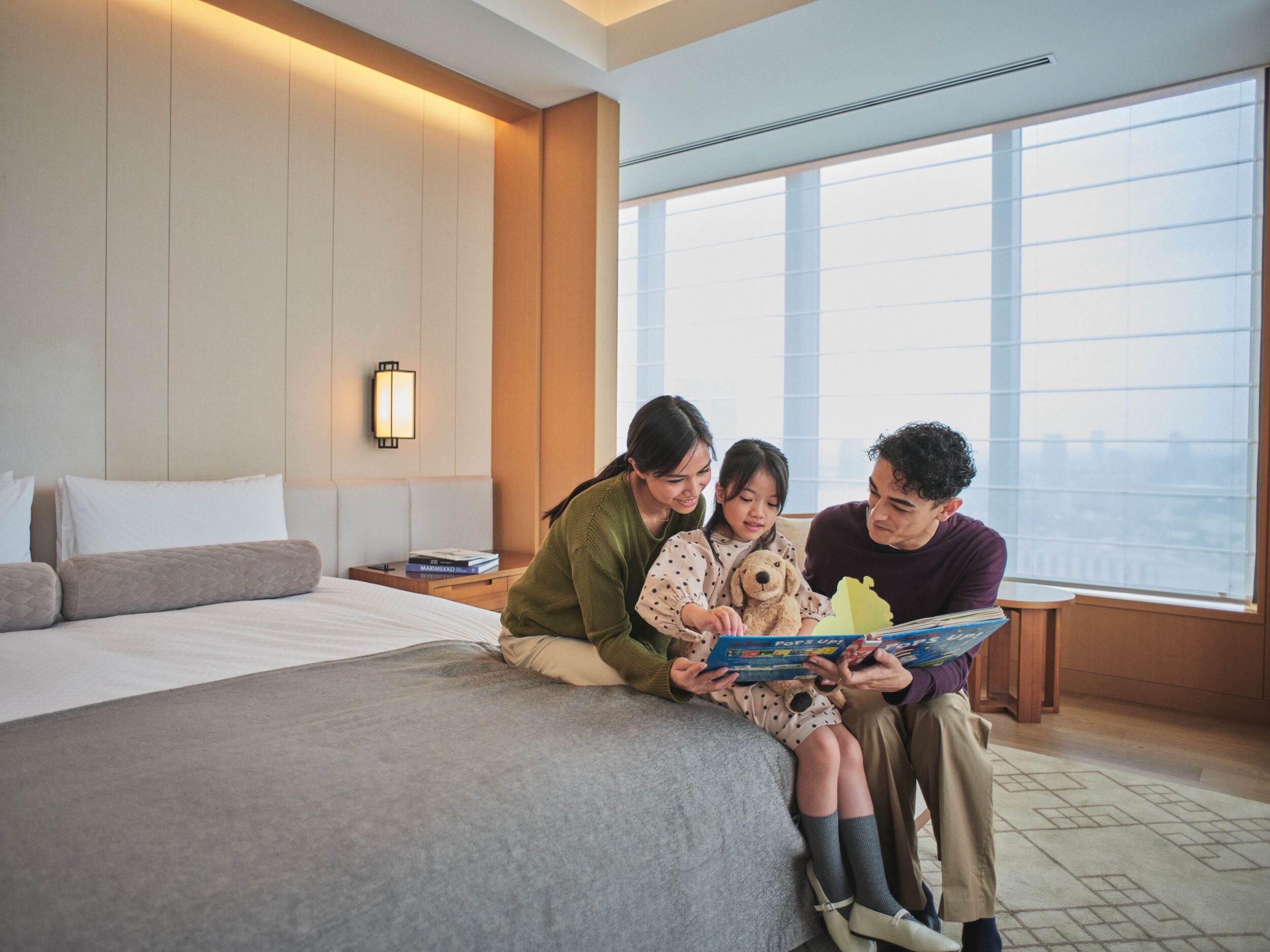 A family—a mother, a father, and a young daughter holding a stuffed animal—sits on a bed in a bright, modern hotel room, reading a picture book together. A large window overlooks a cityscape.