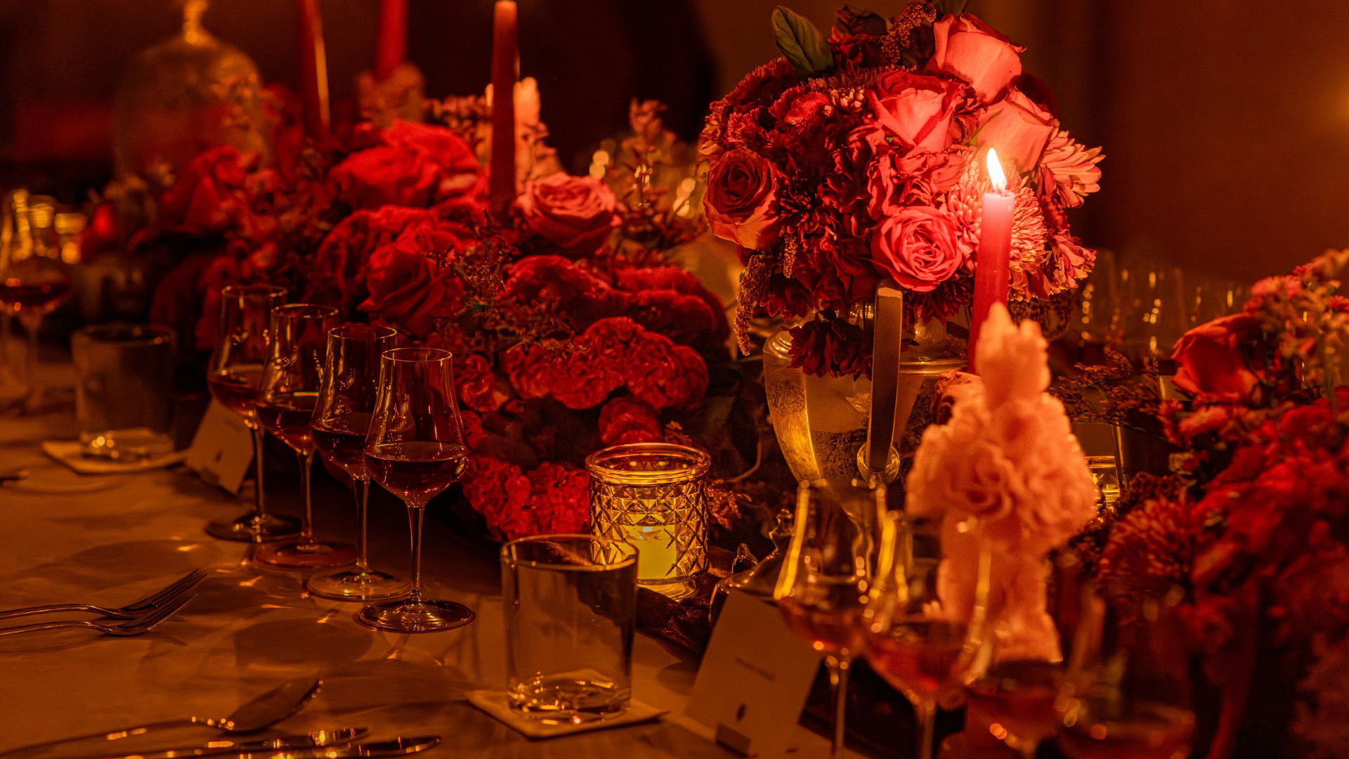 A banquet table with lit candles, bouquets of red flowers, crystal, and silverware.