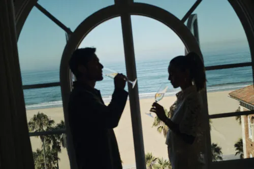 silhouette of 2 people drinking champagne by a window that looks out at the beach