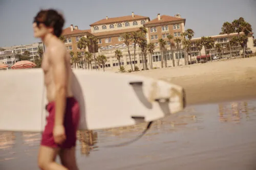 blurry man with a surfboard in the foreground with casa del mar hotel in focus in the background
