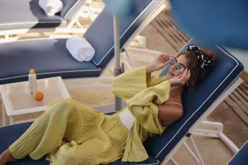 Woman wearing sunglasses and a green outfit relaxing by the pool in a reclining chair.