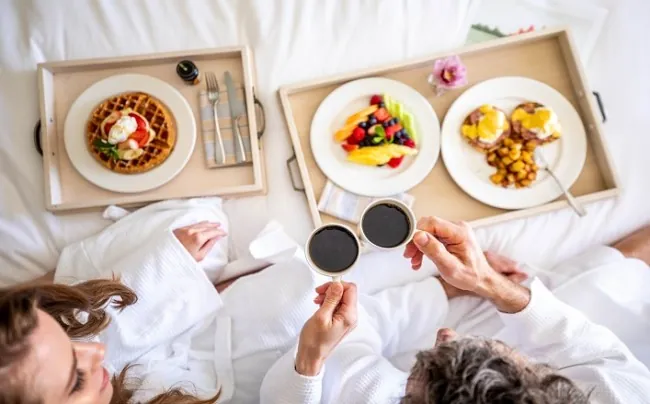 A couple having breakfast in a hotel bed with two cups of black coffee, waffles and fruits.