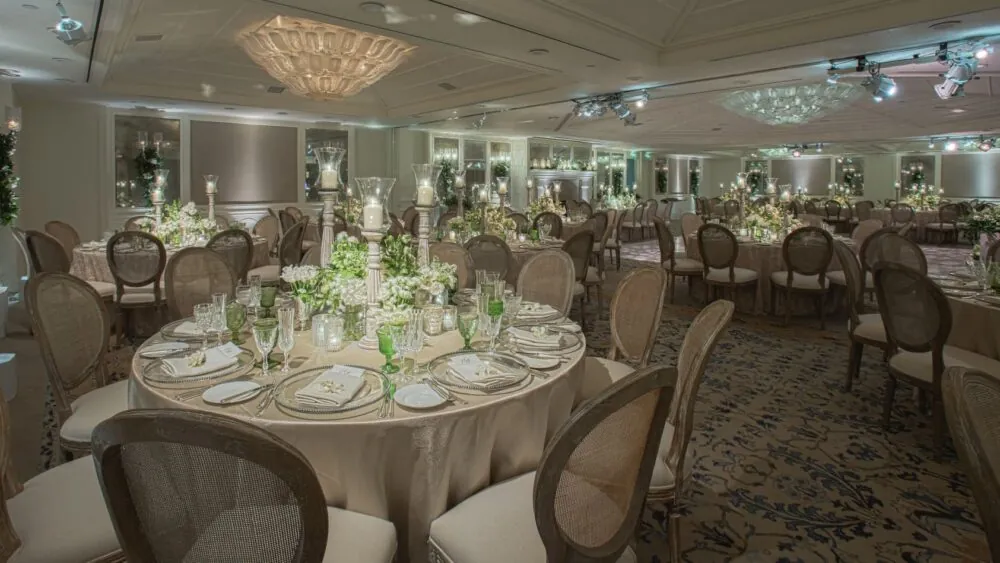 An event venue featuring round tables, chairs, dining setups, & floral arrangements on the tables.