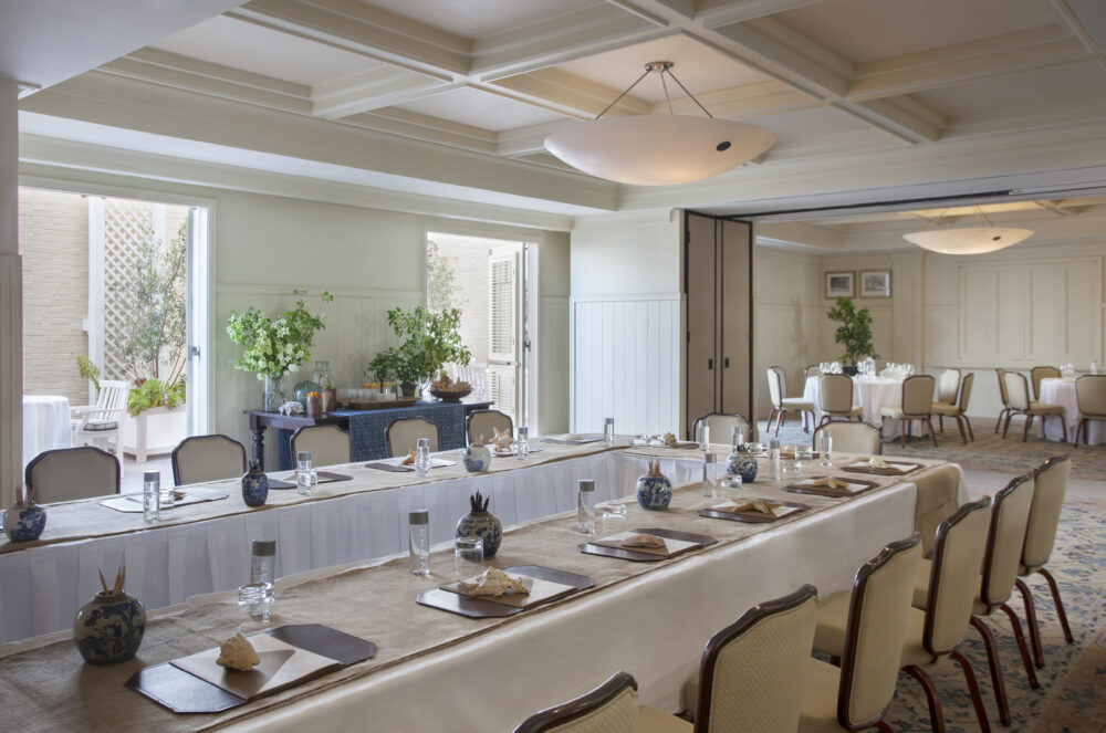 A bright conference room with large tables, each set with water glasses & shells for every chair.
