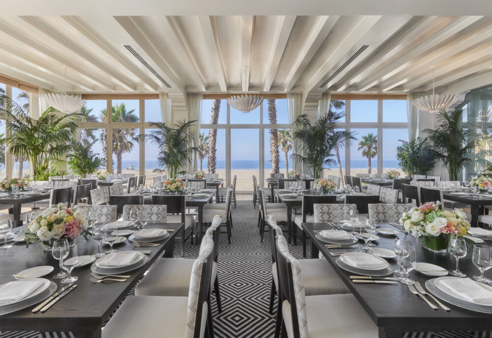 A restaurant featuring square tables, patterned chairs & windows with a view of Santa Monica beach.
