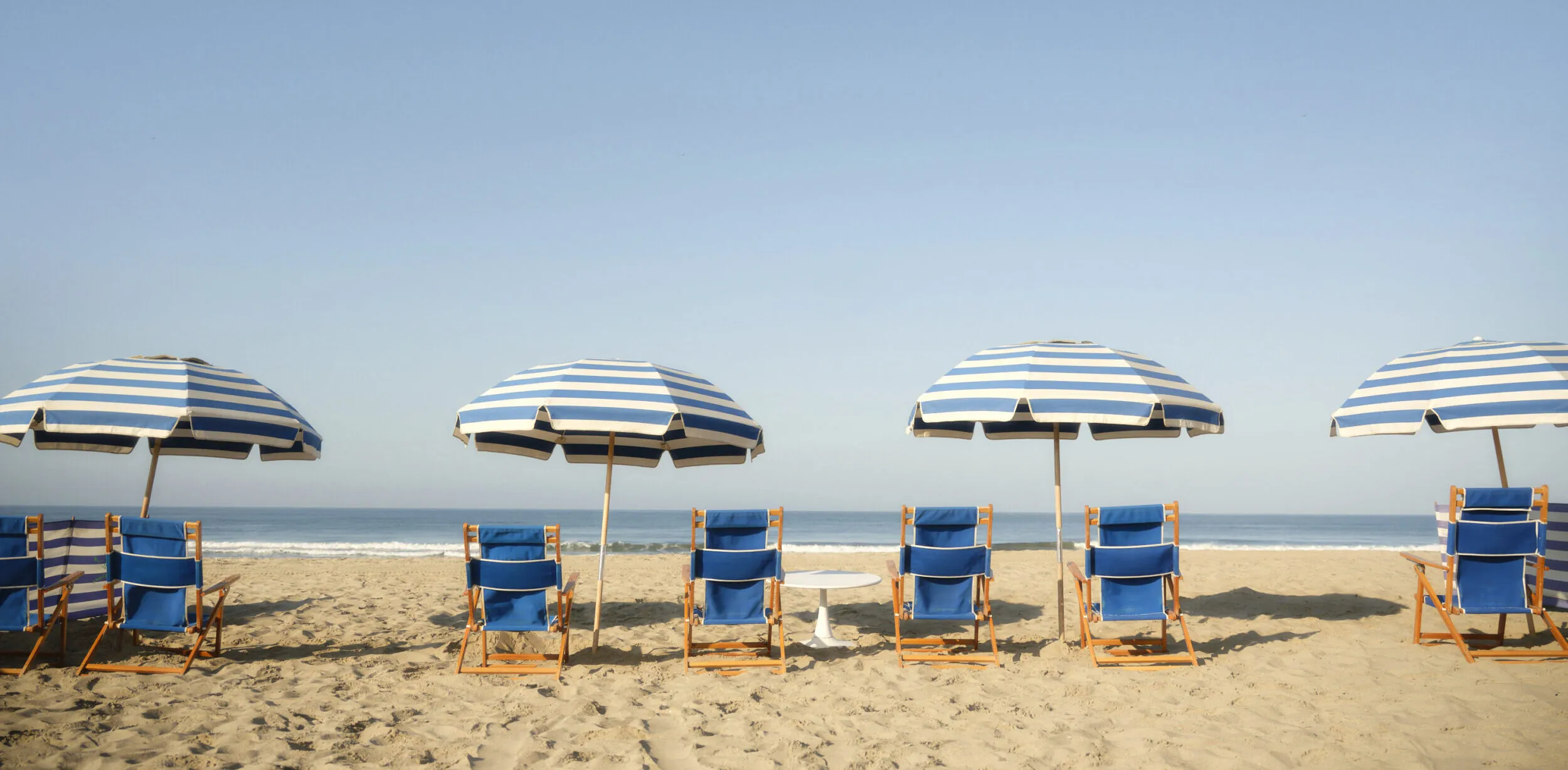 Blue beach chairs sit on the beach under white and blue striped umbrellas.