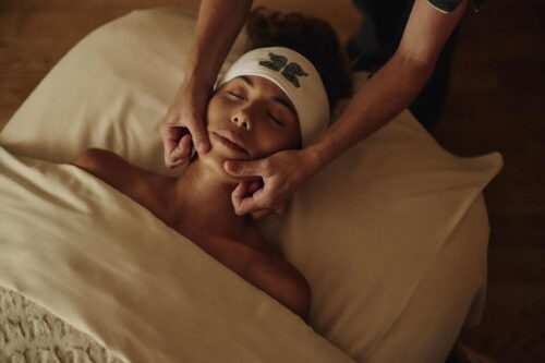 woman laying on spa table, under the sheets, and a masseuse giving her a facial