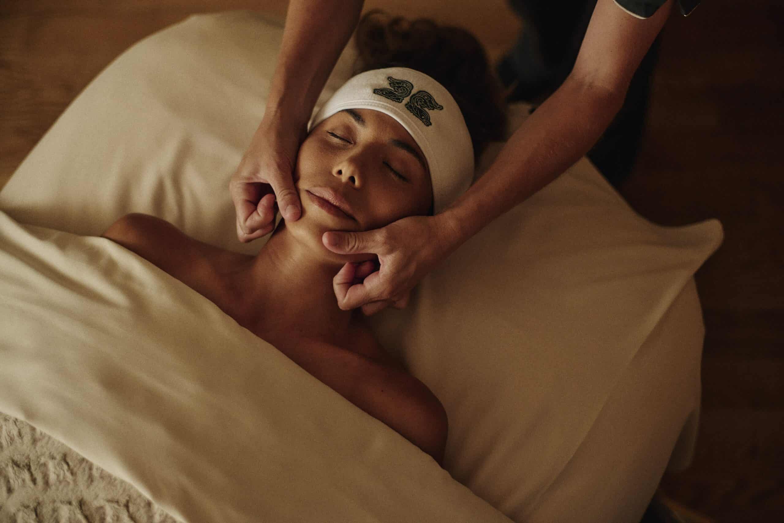 woman laying on spa table, under the sheets, and a masseuse giving her a facial