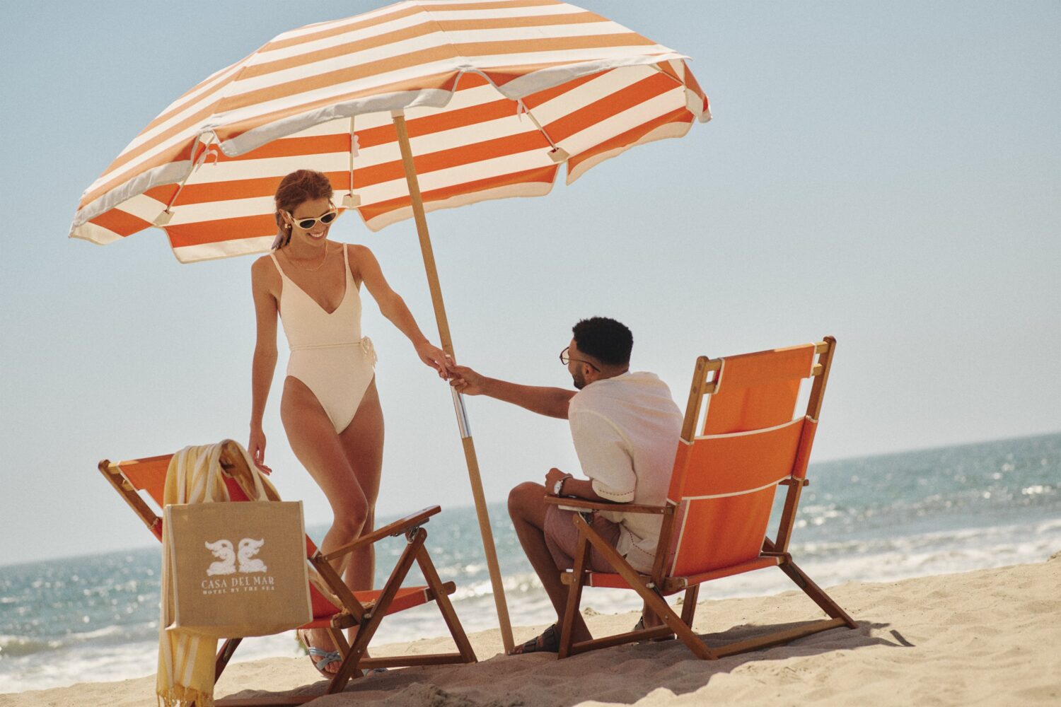 man sitting in a beach chair holding a woman's hand standing in front of him, on the beach with the ocean in background