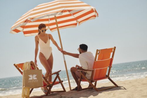 man sitting in a beach chair holding a woman's hand standing in front of him, on the beach with the ocean in background