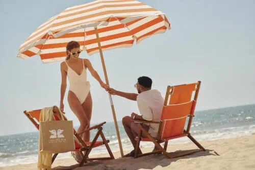 A woman holds a striped beach umbrella while a man sits on an orange beach chair in Santa Monica.