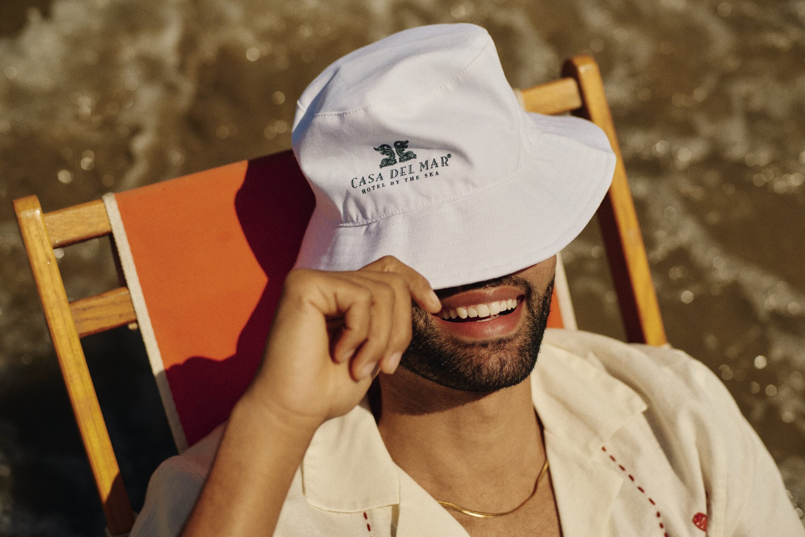 man on the beach, sitting in a beach and wearing a Casa del Mar bucket hat covering his eyes and he is smiling