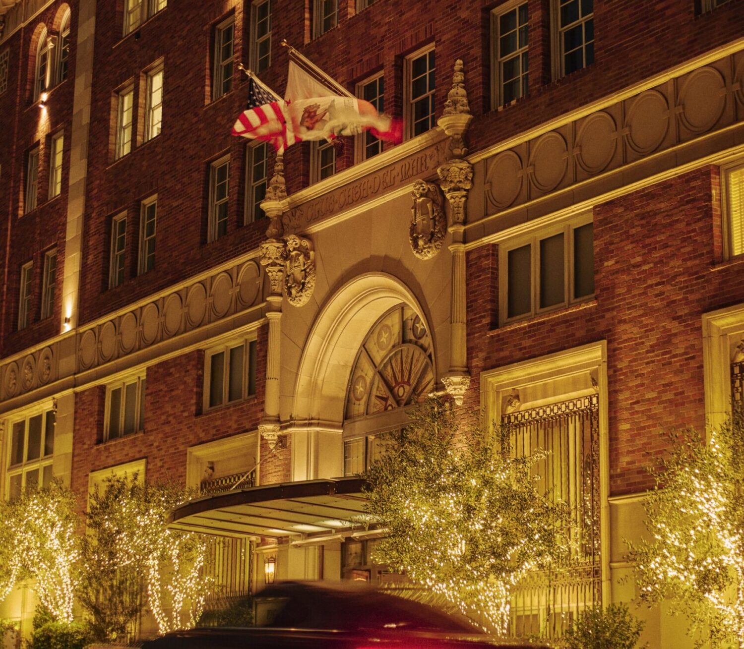 An exterior nighttime shot of Casa del Mar Hotel's front entrance, with lit up trees, flags and a car going by
